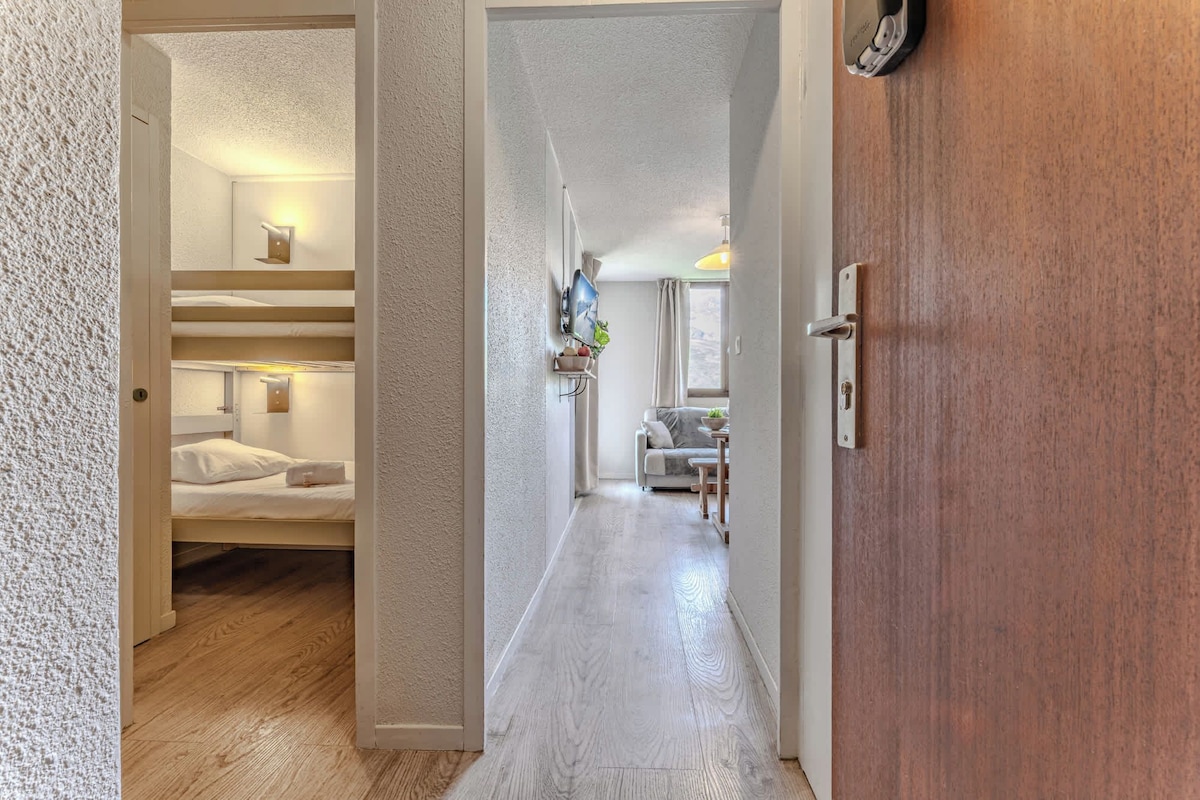 A hotel corridor with a wooden floor, a bunk bed room on the left, a seating area with a table and chairs, and a wooden door on the right.