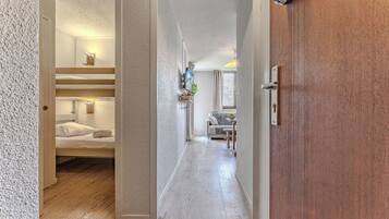 Un pasillo de hotel con suelo de madera, una habitación con literas a la izquierda, un área de estar con una mesa y sillas, y una puerta de madera a la derecha.