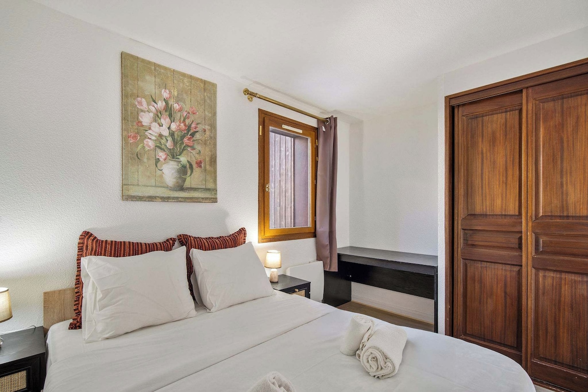 A neatly made bed with white linens and pillows, a bedside table with a lamp, a wooden wardrobe, and a painting of flowers on the wall.