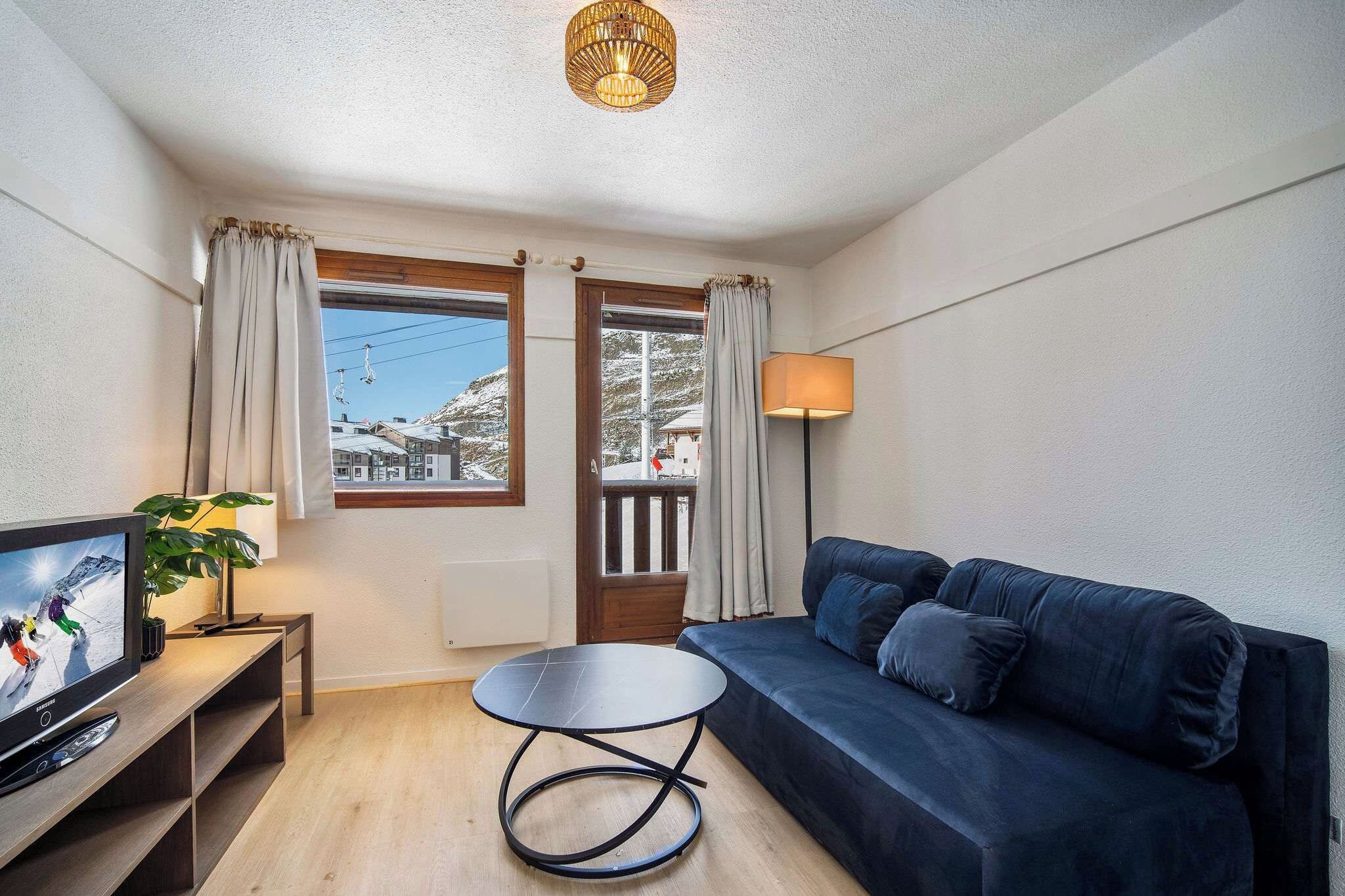 Una sala de estar moderna con un sofá azul oscuro, una mesita redonda pequeña, un televisor sobre un mueble de madera, una lámpara y vistas a una montaña nevada a través de una ventana.