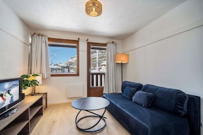 A modern living room with a dark blue sofa, a small round table, a TV on a wooden stand, a lamp, and a view of a snowy mountain through a window. - AEA - Les Belleville (Les Belleville,)