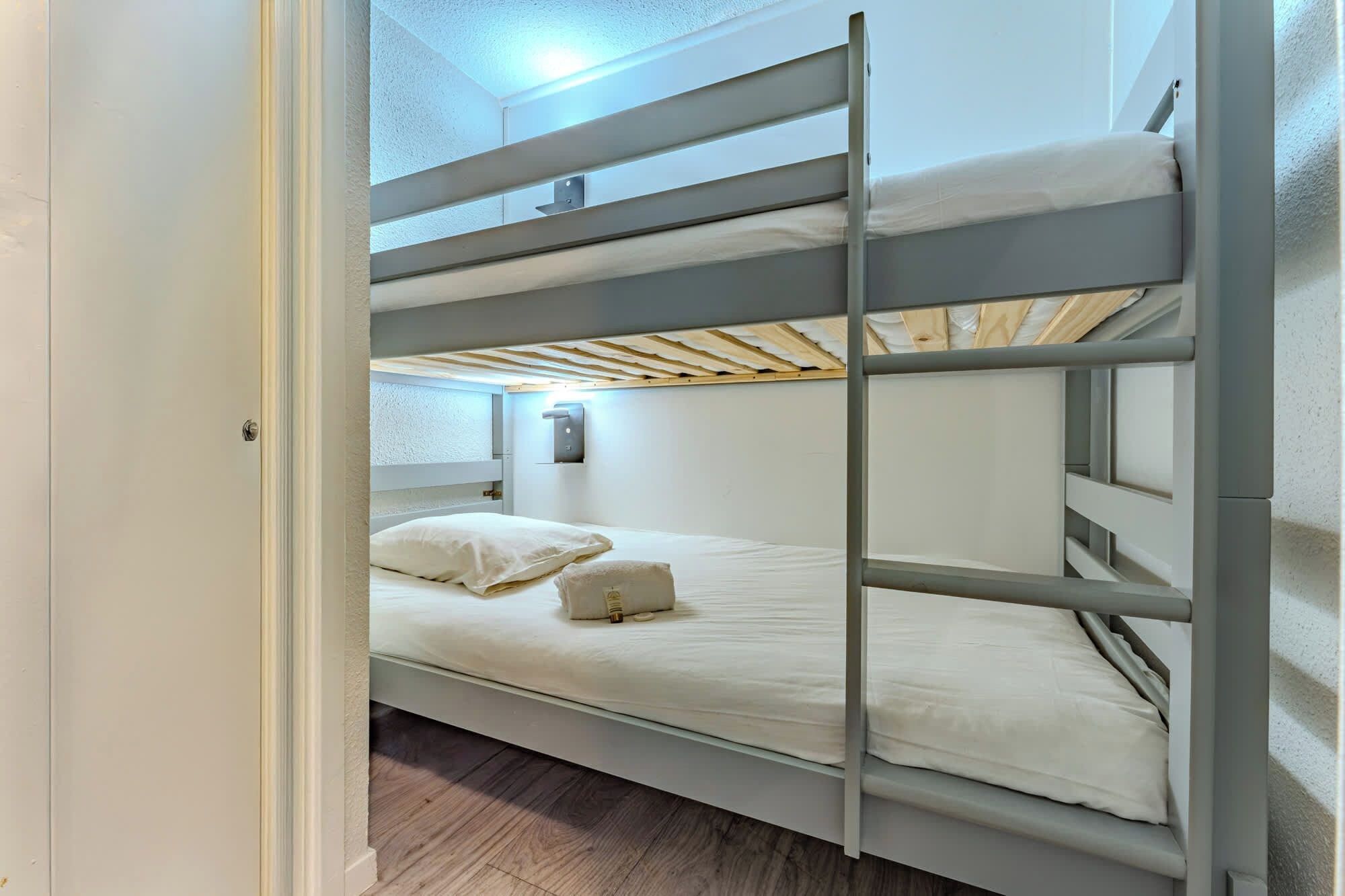 A bunk bed in a room with a wooden floor, white walls, and a small bedside table with a lamp.