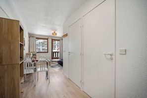 A compact room with a wooden wardrobe, a dining table with chairs, a small round table, and a door leading to an outdoor area.