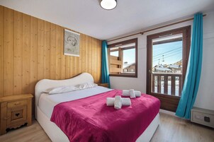A bedroom with a wooden wall, a bed with a pink blanket, a nightstand, and a window with a view of buildings.