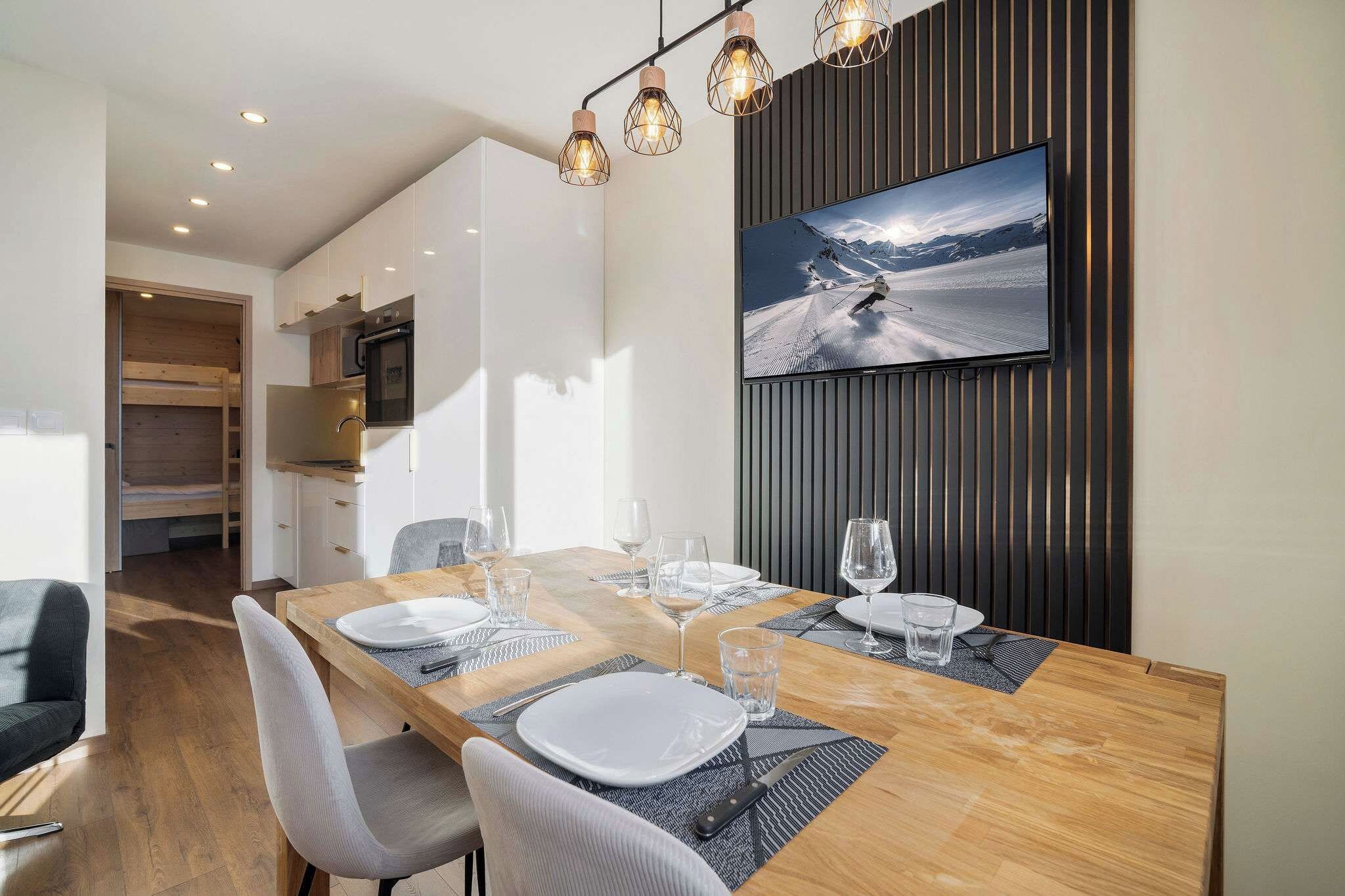 A modern dining area with a wooden table set for four, a mounted flat-screen TV, and a kitchen in the background.