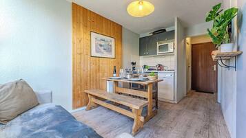 A cozy living room with a wooden bench, a dining table set for two, a kitchen with a microwave, and a door leading to another room.