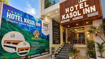 Entrada de hotel con un cartel publicitando servicio de habitaciones las 24 horas, habitaciones de lujo, ideal para parejas, estadía perfecta para negocios y familias, servicio al huésped 24 horas, estacionamiento y WiFi gratuito.