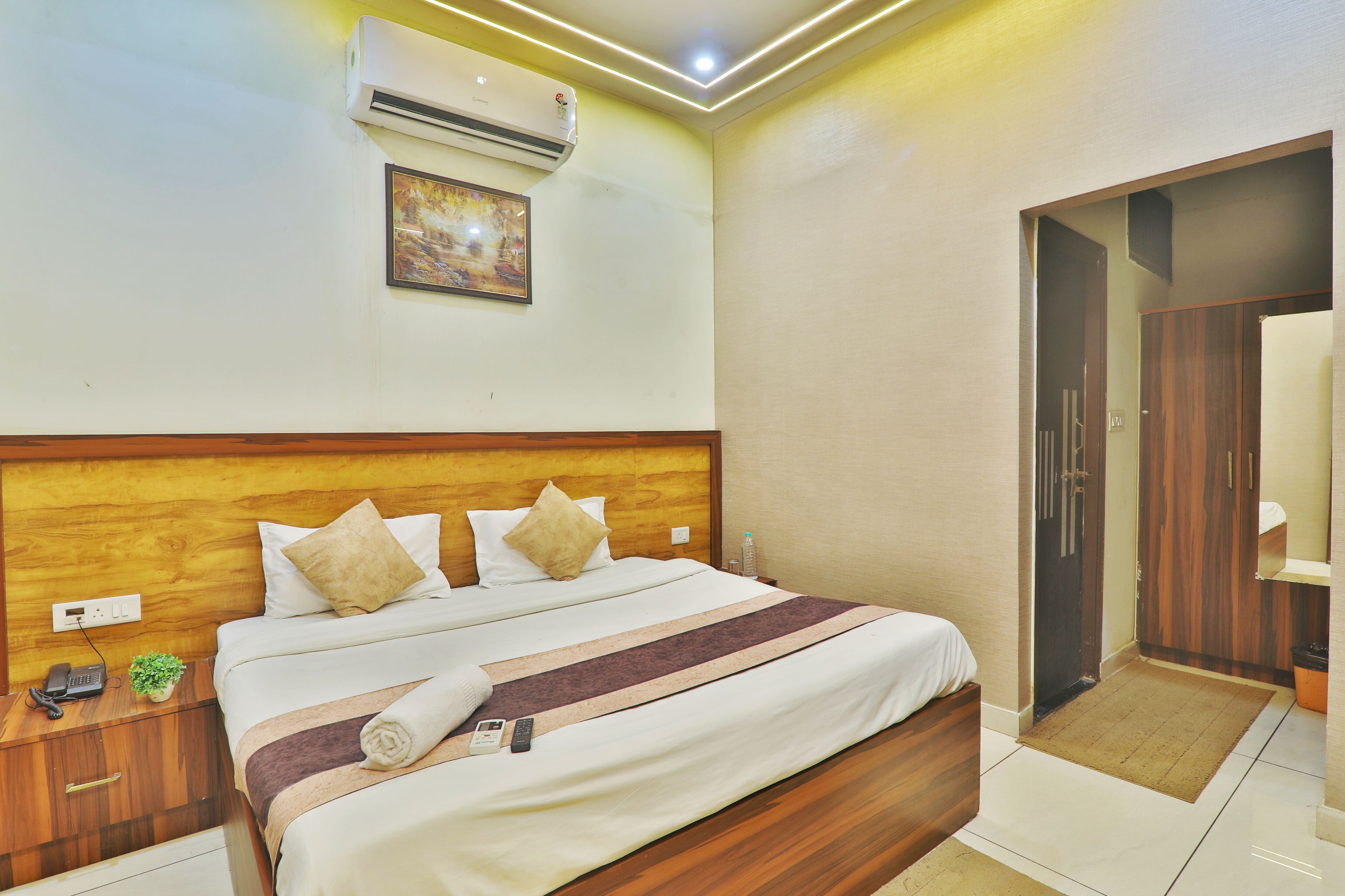 A hotel room with a wooden headboard, a bed with white bedding and a striped blanket, a bedside table with a phone and a plant, and an air conditioning unit on the wall.