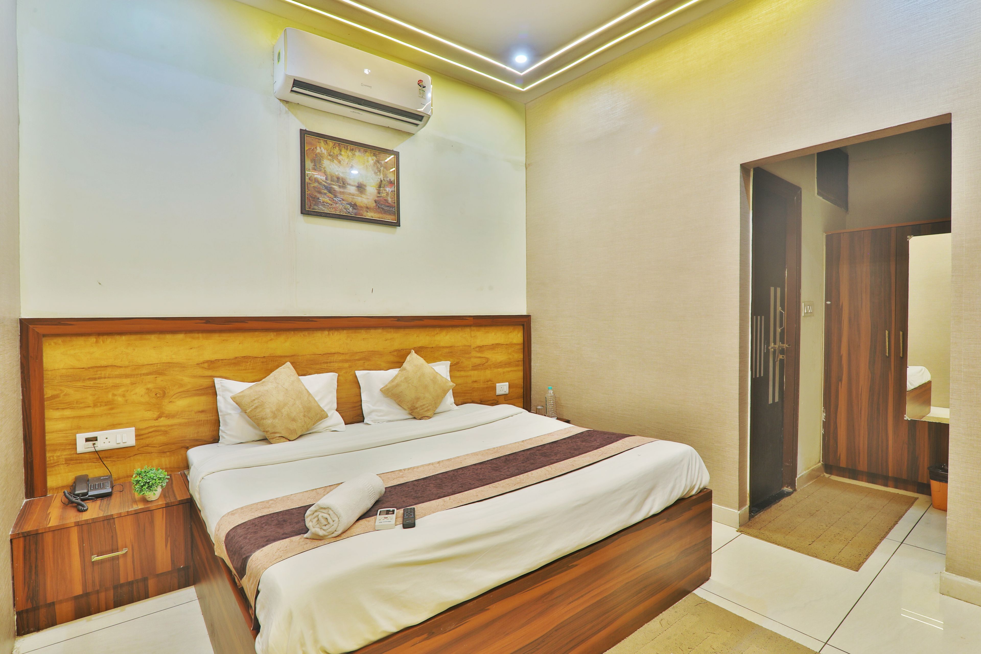 A hotel room with a wooden headboard, a bed with white bedding and a striped blanket, an air conditioning unit, a framed picture on the wall, and a wooden wardrobe.