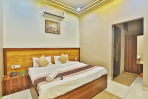 A hotel room with a wooden headboard, a bed with white bedding and a striped blanket, an air conditioning unit, a framed picture on the wall, and a wooden wardrobe. - Hotel Kasol Inn (Gorakhpur)