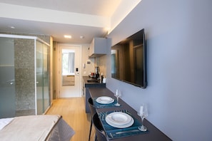 A hotel room with a bed, a dining area with a table set for two, a flat-screen TV mounted on the wall, and a bathroom with a glass shower enclosure. - Setai Yacht - Flat in Cabo Branco by Carpediem (João Pessoa)