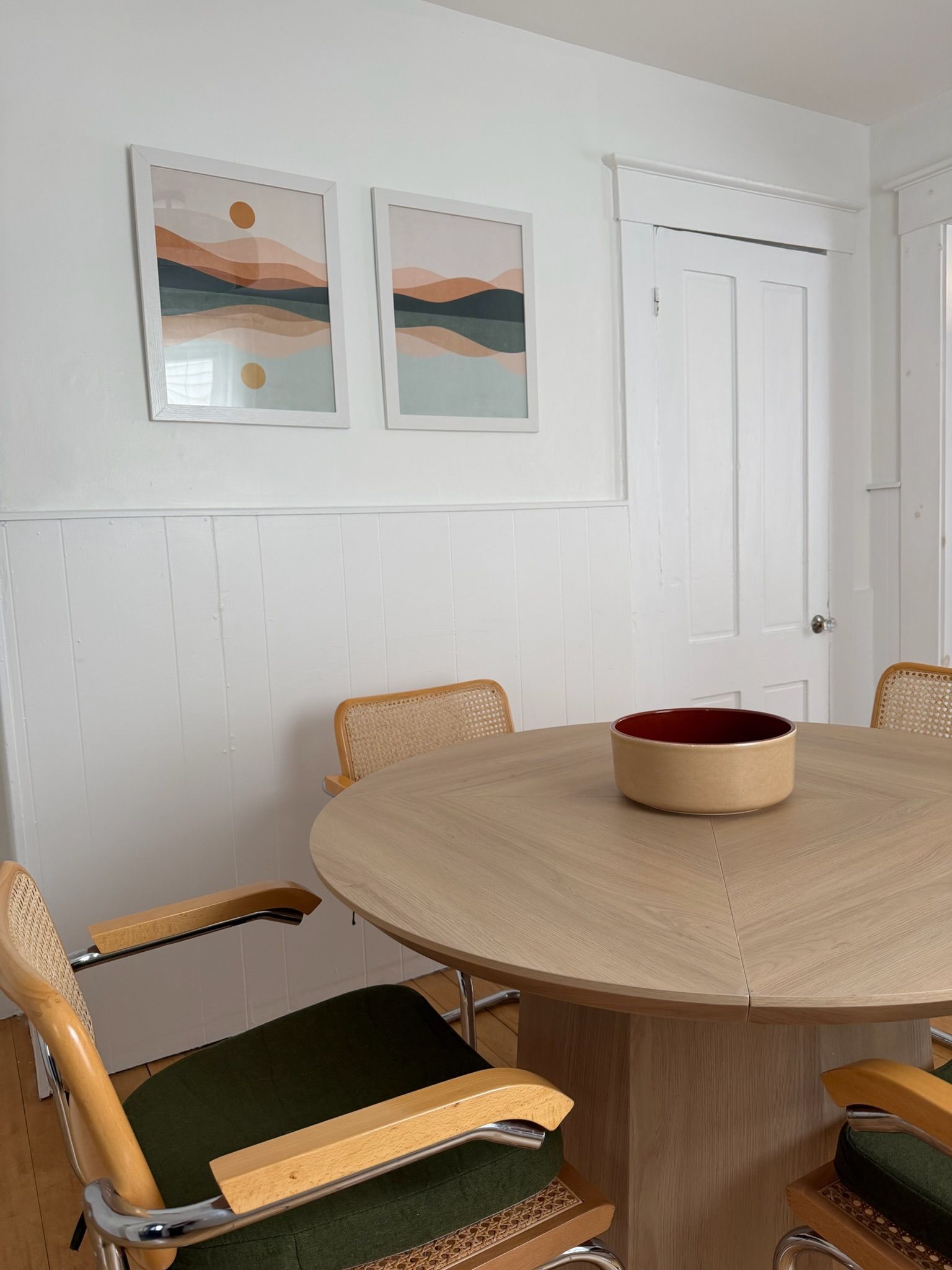 Een eetgedeelte met een ronde tafel, stoelen en twee ingelijste kunstwerken aan de wand.
