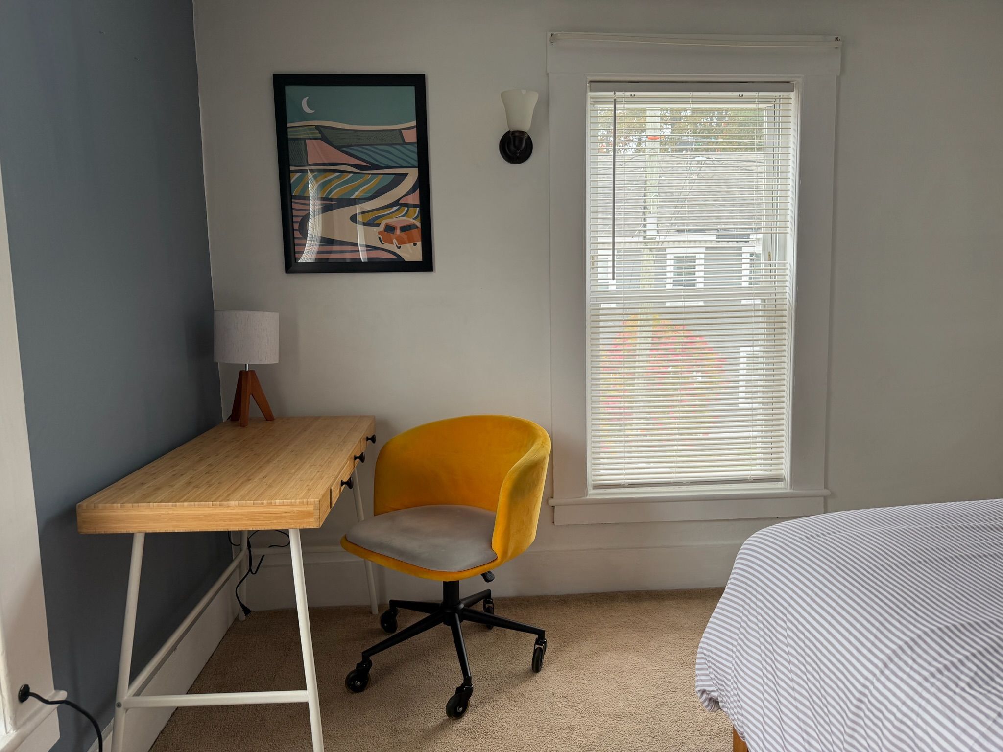 A bedroom with a bed, a desk, a chair, a window with blinds, and a wall-mounted lamp.