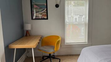 A bedroom with a bed, a desk, a chair, a window with blinds, and a wall-mounted lamp.