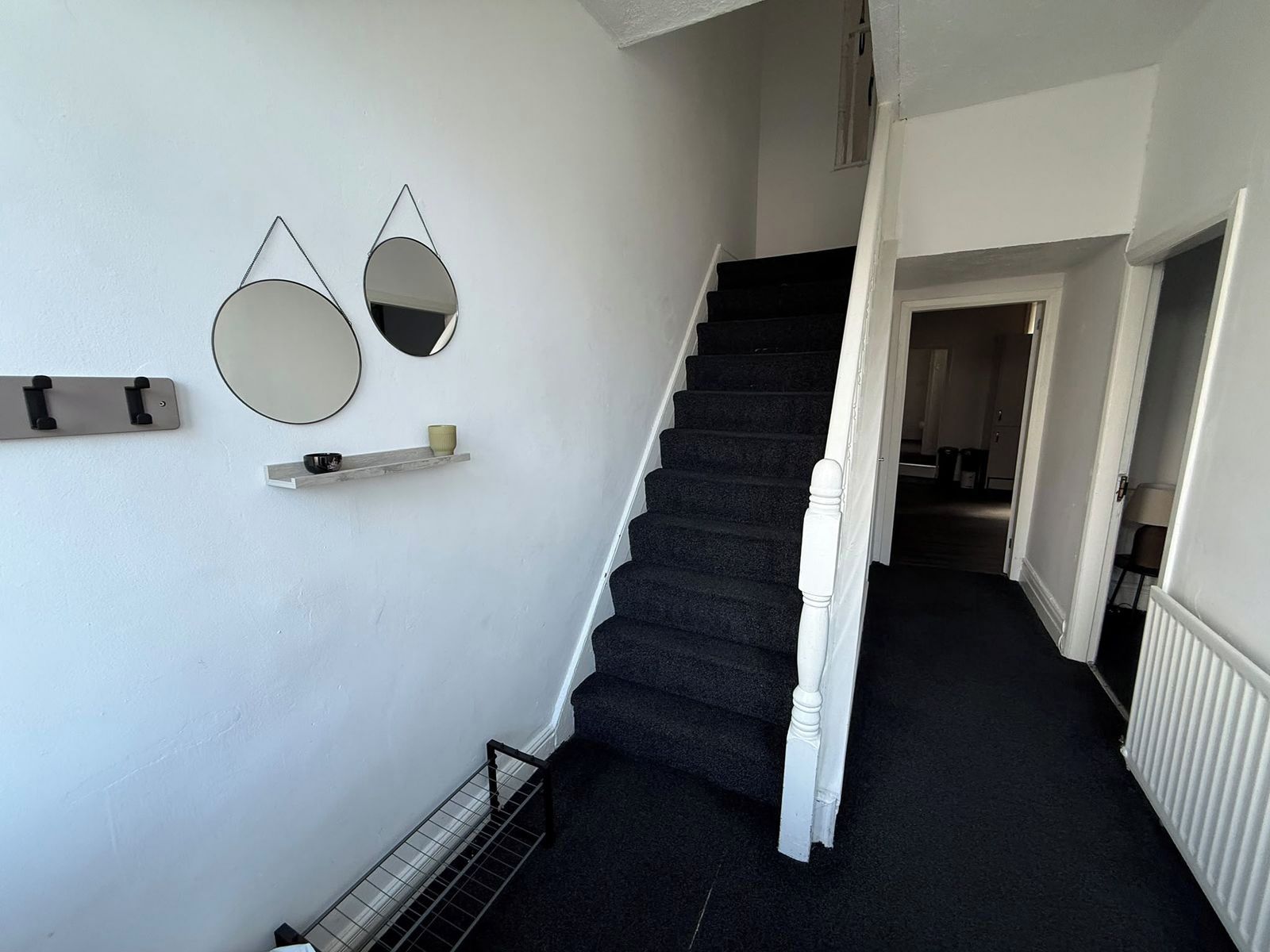 A staircase with a black carpet leading to a landing with a white wall. There are two wall-mounted mirrors and a shelf with a cup.