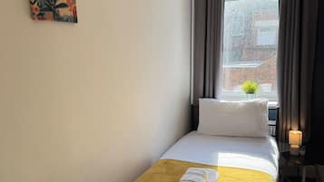 Een kleine eenpersoonskamer met een raam, een nachtkastje met een lamp en twee kunstwerken aan de muur.