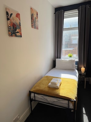 Et lille værelse med en enkeltseng, et vindue, et natbord med en lampe og to billeder hængt på væggen.