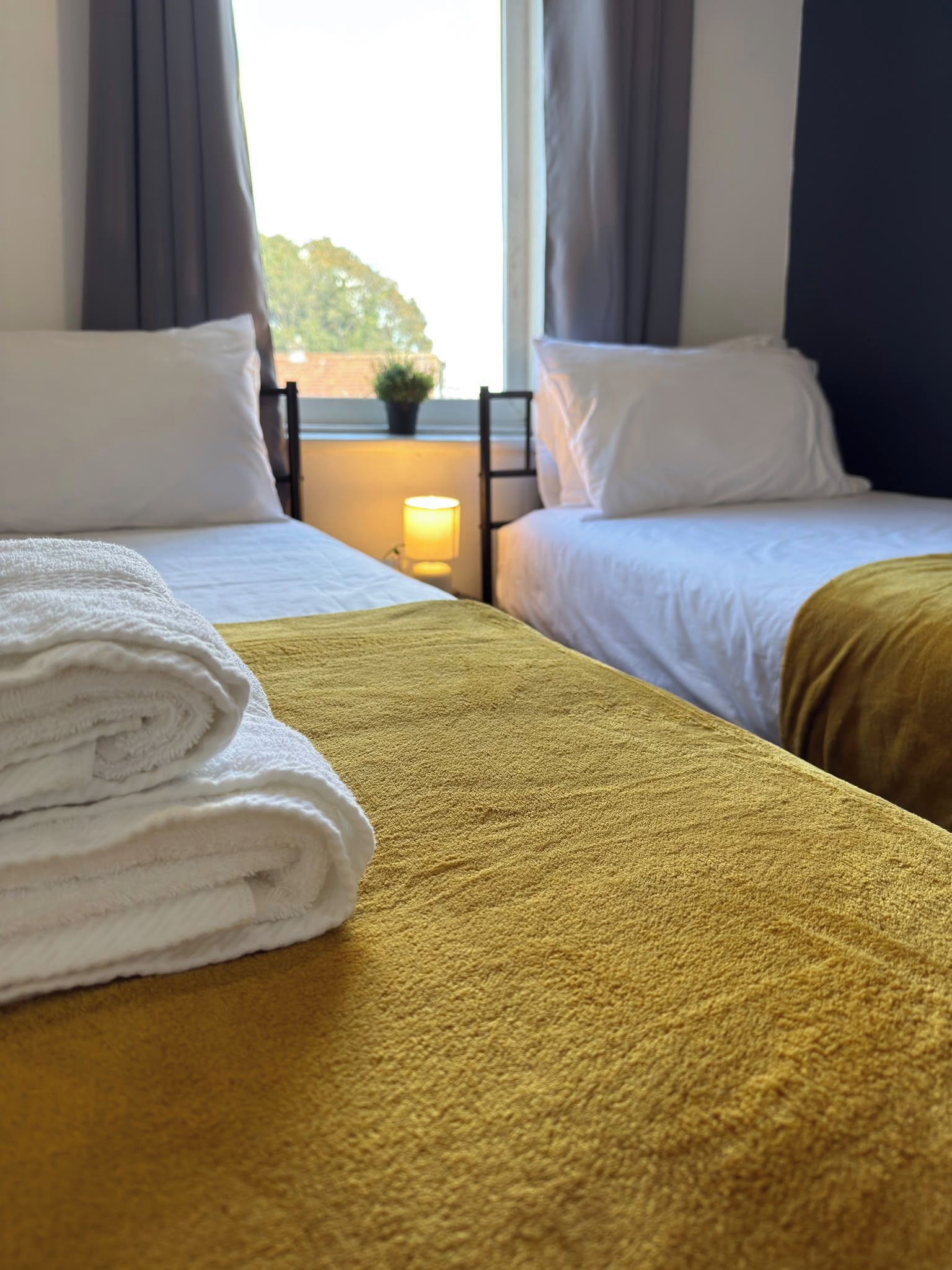 A hotel room with two beds, a window with curtains, a bedside lamp, and a view of greenery outside.