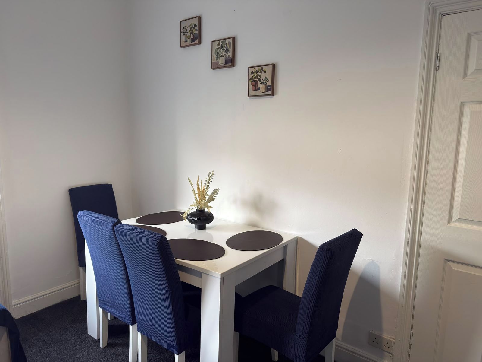 A dining area with a white table, four blue chairs, and a vase with dried plants.