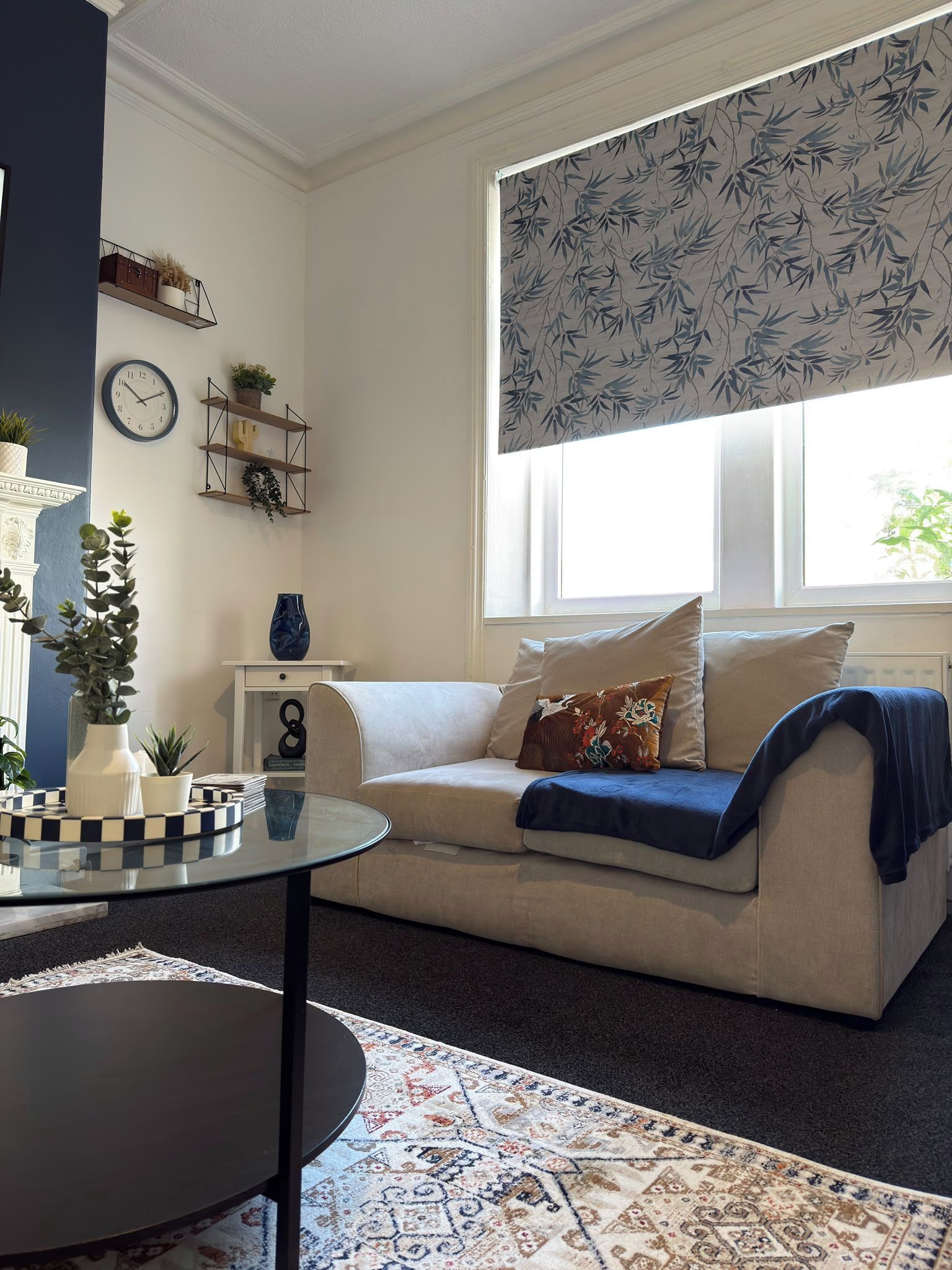 A living room with a sofa, a clock, shelves with plants, and a window with a bamboo pattern.