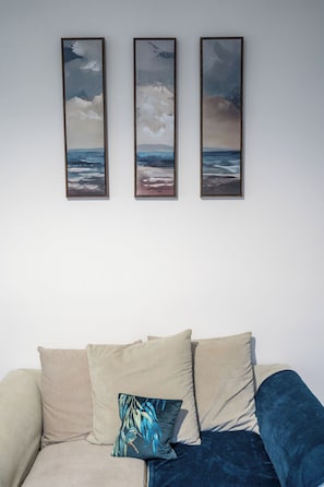 Et værelse med en sofa, tre billeder i rammer der viser kystmotiver, og en pude med et fuglemotiv.