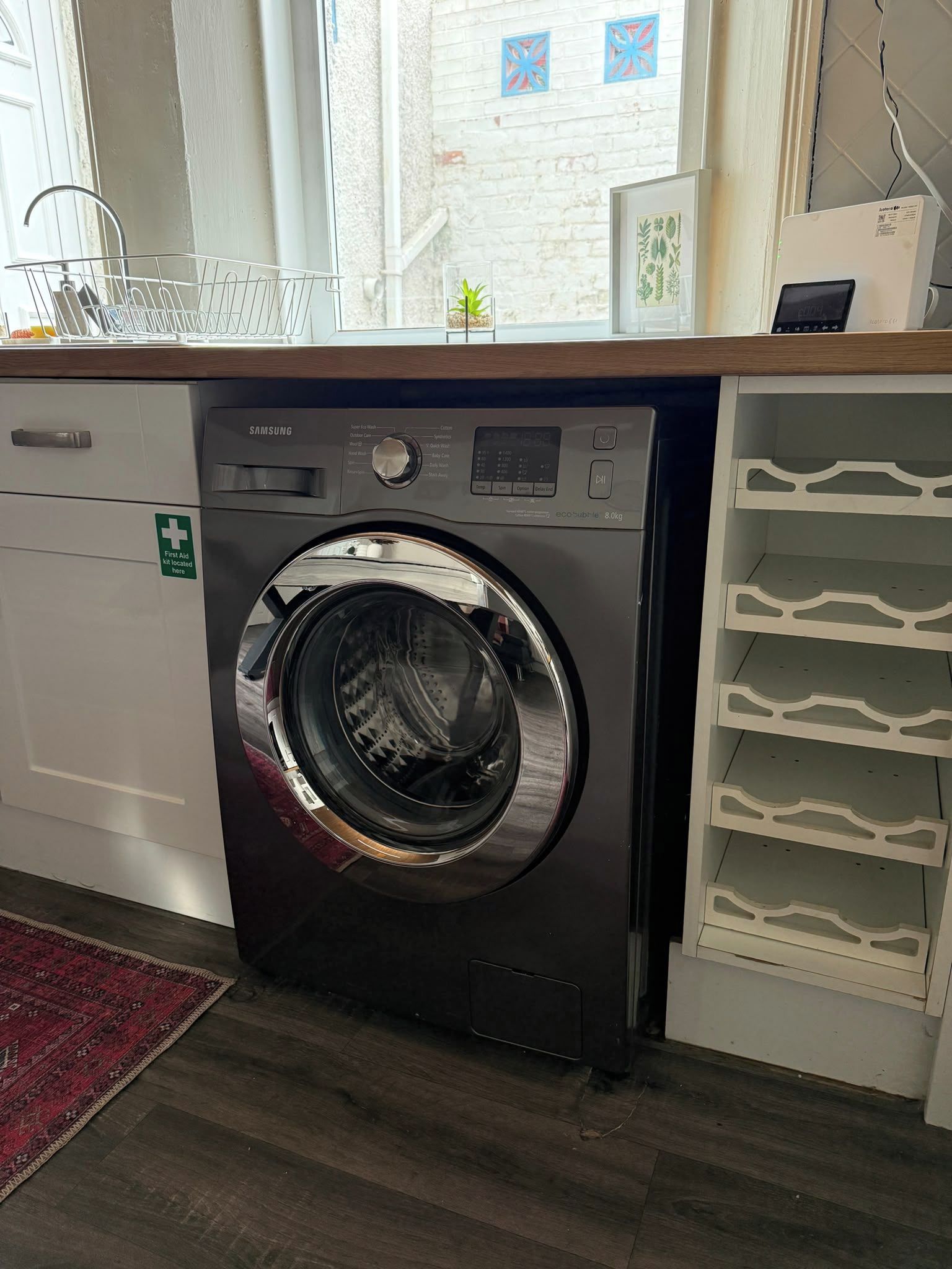 A Samsung front-loading washing machine in a room with a window, a small plant, and a first aid kit sign.