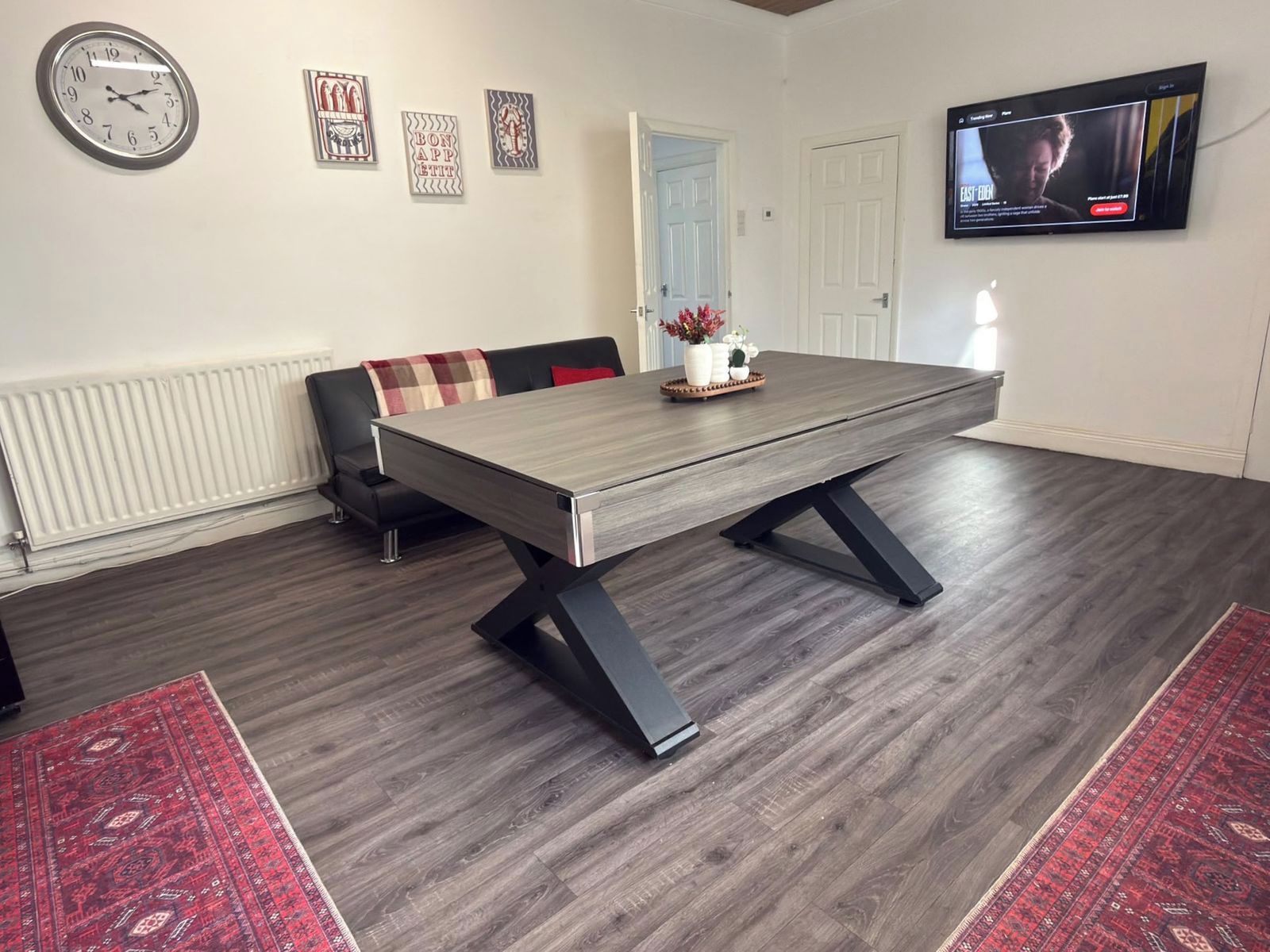 A dining area with a rectangular table, black chairs, a clock, framed artwork, a radiator, a television, and a red patterned rug.
