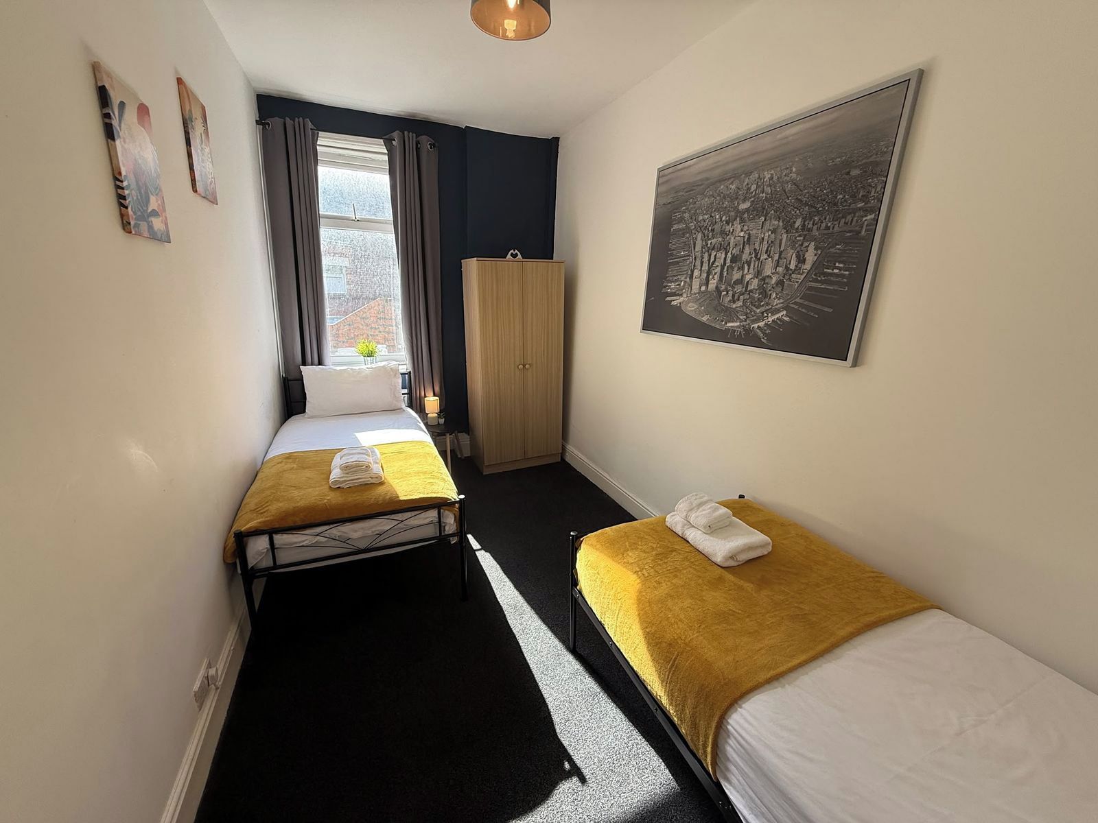 A hotel room with two single beds, a wardrobe, a window with curtains, and a framed cityscape picture on the wall.