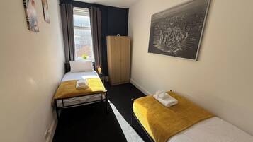 A hotel room with two single beds, a wardrobe, a window with curtains, and a framed cityscape picture on the wall.