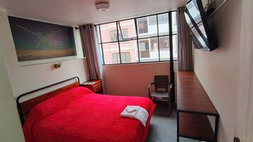 A hotel room with a bed, a desk, a chair, a TV, and a window with a view of buildings.