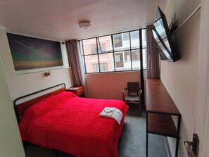 A hotel room with a bed, a desk, a chair, a TV, and a window with a view of buildings. - Hostal Kalopsia Suites (La Paz)
