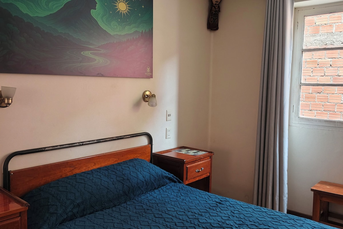 Een kamer met een bed, een nachtkastje, een schilderij aan de muur, een raam met gordijnen en een bakstenen muur.