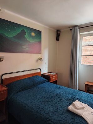 Een kamer met een bed, een nachtkastje, een schilderij aan de muur, een raam met gordijnen en een bakstenen muur.