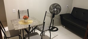 Ein kleiner Wohnbereich mit Esstisch, Stühlen, einem schwarzen Sofa und einem Standventilator.