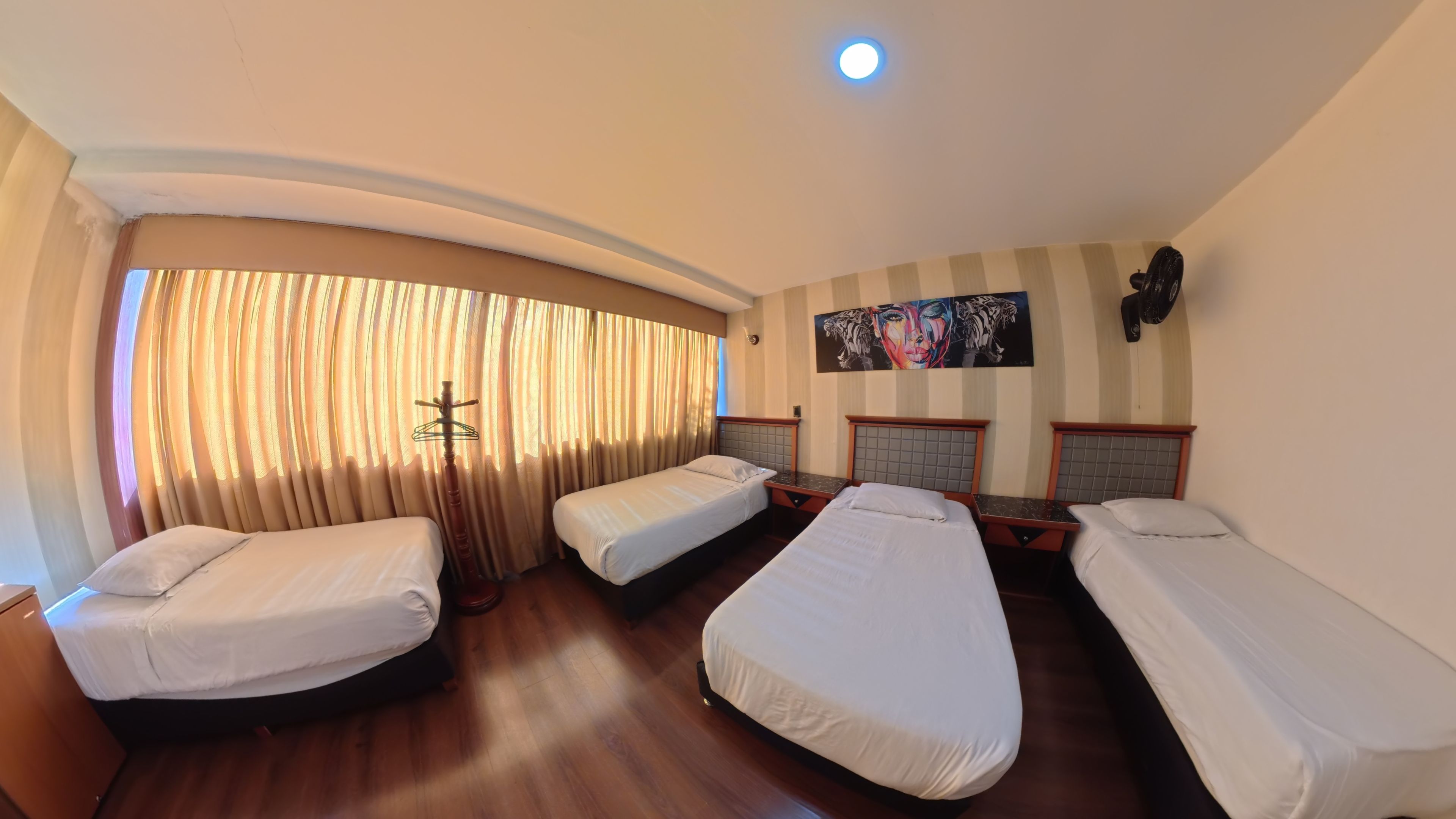Ein Hotelzimmer mit vier Einzelbetten, Holzboden, großem Fenster mit Vorhängen, einem Bild an der Wand und einer Deckenleuchte.