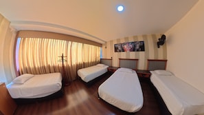 Quarto de hotel com quatro camas de solteiro, piso de madeira, uma janela grande com cortinas, um quadro na parede e uma luminária no teto.