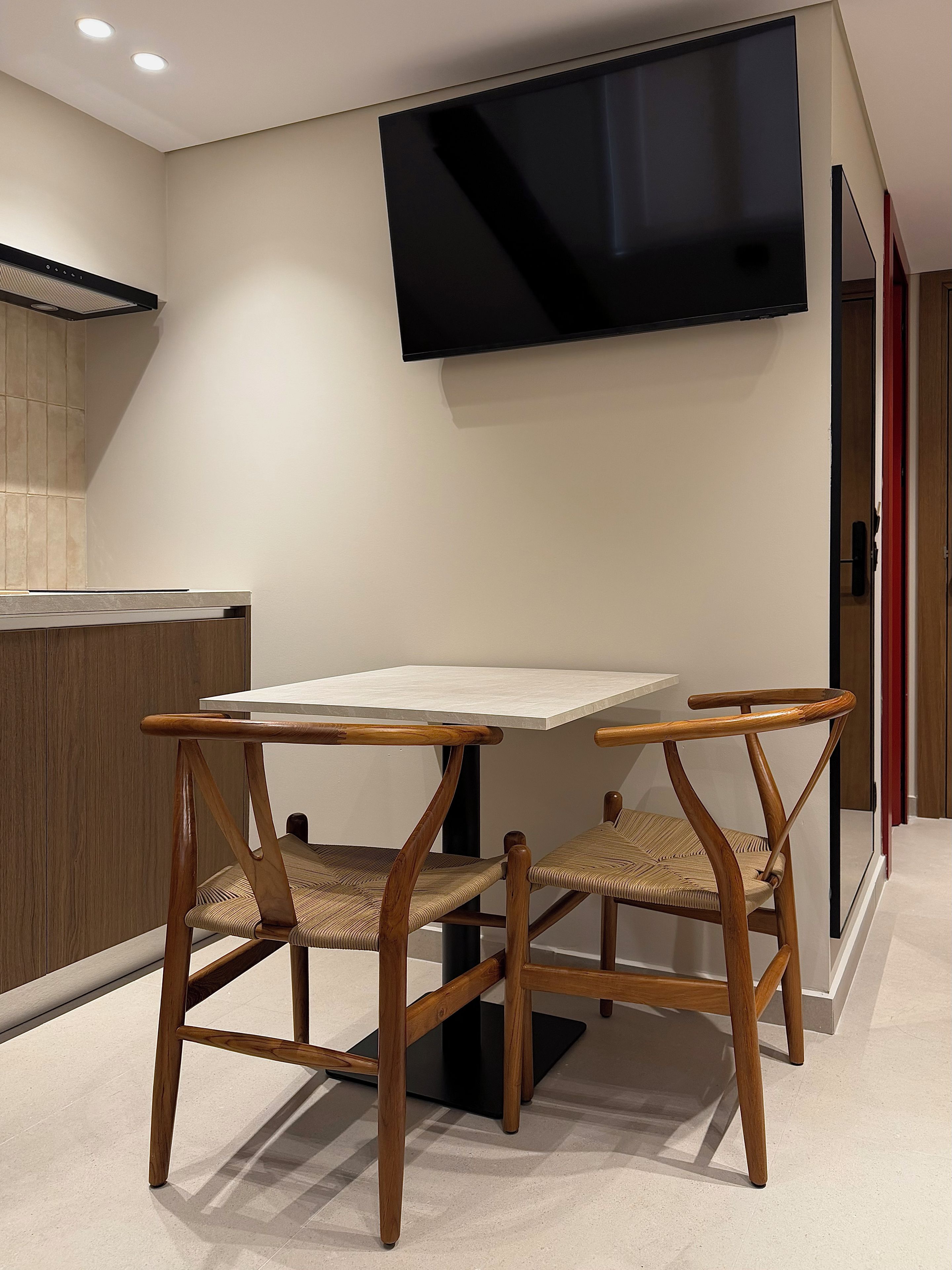 A modern kitchen with a flat-screen TV mounted on the wall, a white countertop, and two wooden chairs with woven seats.