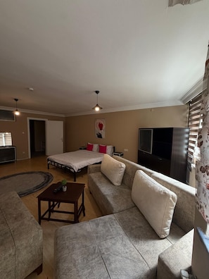 A living room with a sofa, a coffee table, a bed with pillows, a TV, and a window with blinds. - Cozy 1-bedroom Apartment in Pendik, Türkiye (Istanbul)