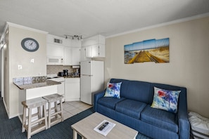 Ein kompakter Wohnraum mit einer Küchenzeile, einem blauen Sofa mit bunten Kissen und einem Landschaftsbild an der Wand.