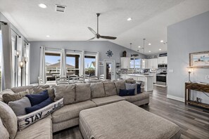 A spacious living room with a large sectional sofa, a ceiling fan, and a view to the outside. - Salty Pelican | Easy Beach Access | Fenced Yard (Bolivar Peninsula)