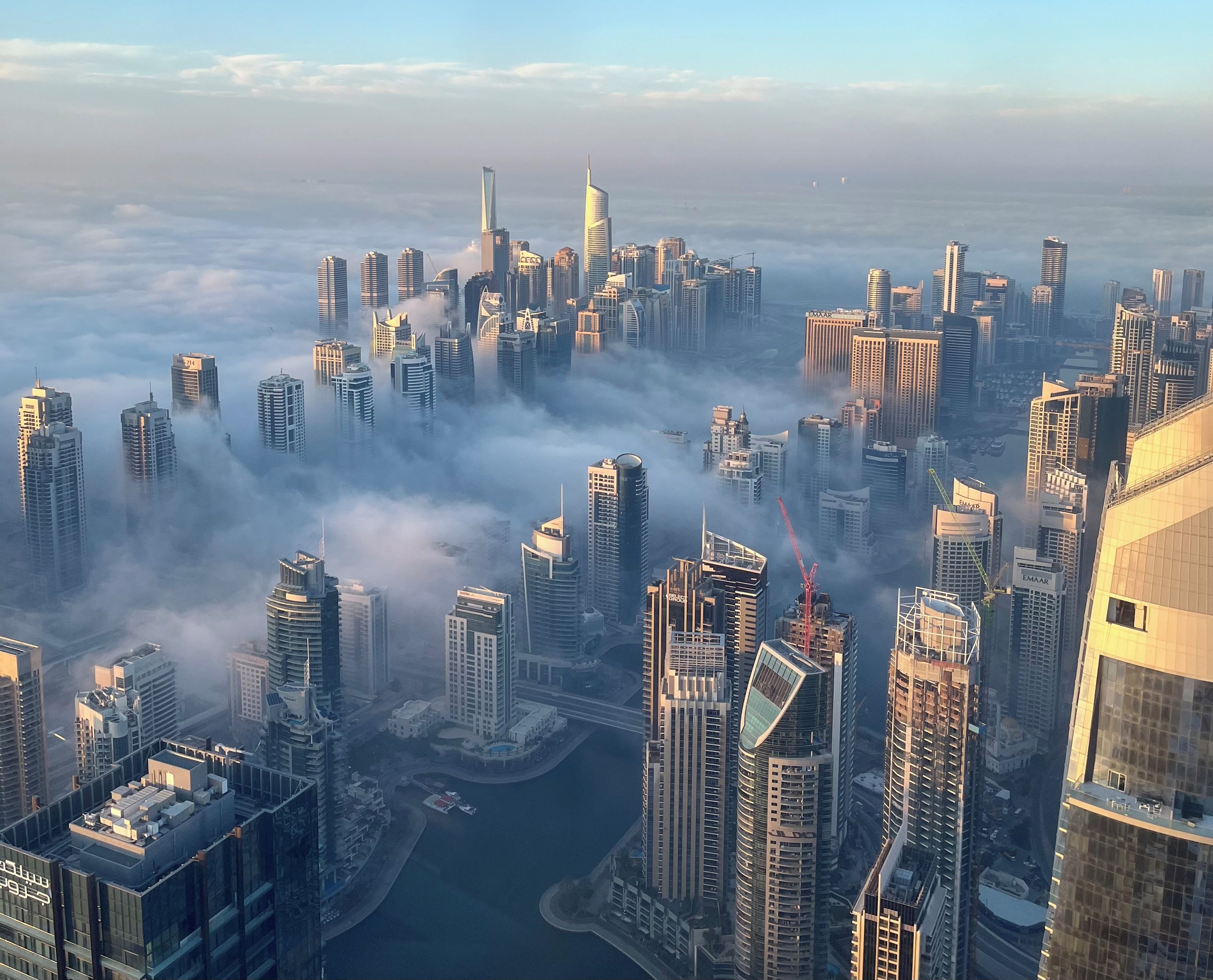 A high-rise urban area with multiple skyscrapers, a body of water, and a dense cloud cover.