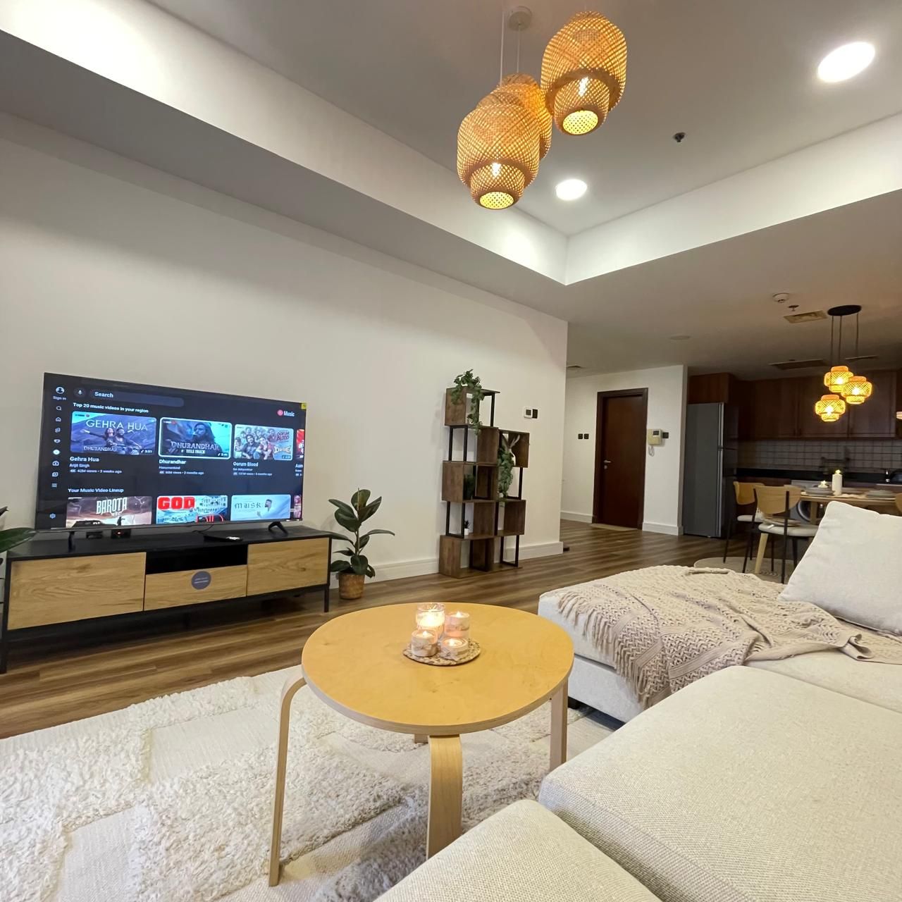 Un salón moderno con televisor de pantalla plana, muebles de madera, una mesa redonda de centro con velas y una cocina al fondo.