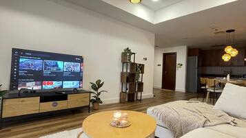 Un salón moderno con televisor de pantalla plana, muebles de madera, una mesa redonda de centro con velas y una cocina al fondo.