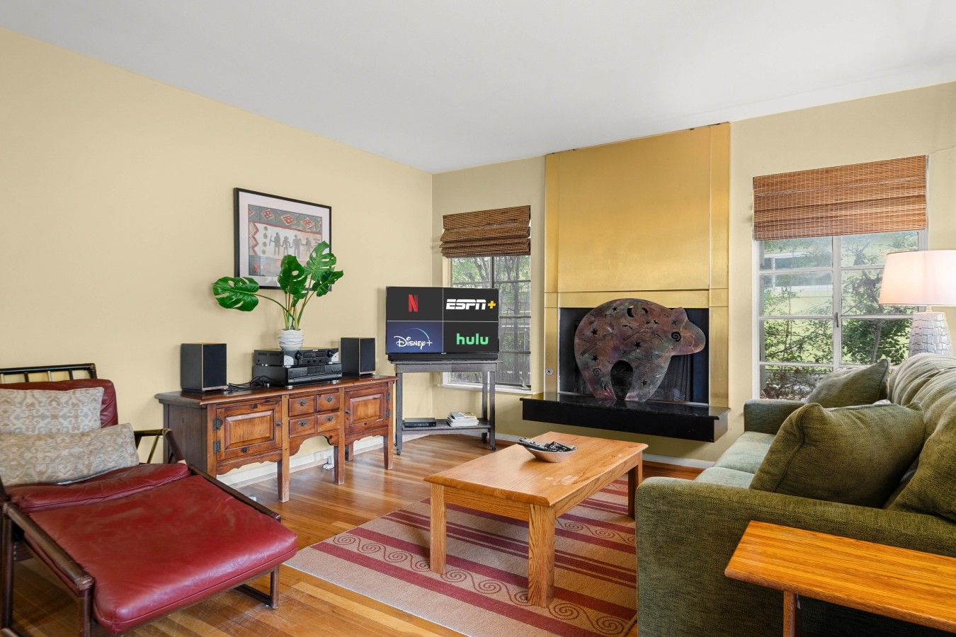 Een woonkamer met een bank, een rode fauteuil, een houten salontafel, een televisie met logo's van streamingdiensten, een open haard met een decoratief beeldhouwwerk van een dier, en een raam met uitzicht.