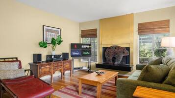 Een woonkamer met een bank, een rode fauteuil, een houten salontafel, een televisie met logo's van streamingdiensten, een open haard met een decoratief beeldhouwwerk van een dier, en een raam met uitzicht.