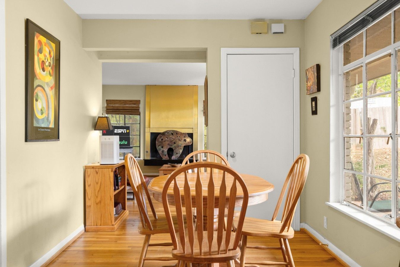 A dining area with a round table and wooden chairs, a fireplace, and a view through a window.