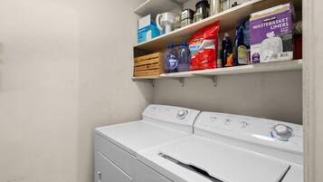 A laundry area with a front-loading washer and dryer, shelves above containing cleaning supplies, and a metal container on the shelf.