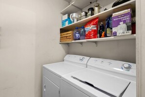 A laundry area with a front-loading washer and dryer, shelves above containing cleaning supplies, and a metal container on the shelf. - Eclectic South Austin Home w Flex Studio Room & Covered Patio (Austin)