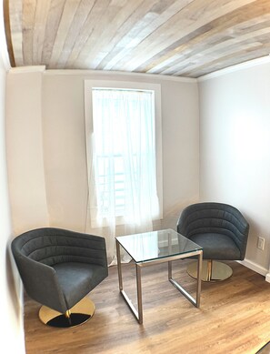 Une pièce comprenant deux fauteuils, une table avec un plateau en verre, une fenêtre ornée de rideaux légers et un plafond en bois.