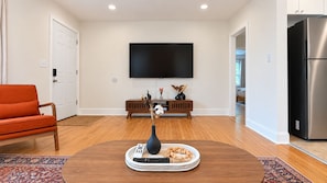Ein Wohnzimmer mit einem an der Wand befestigten Flachbildfernseher, einem Holztisch mit einer Vase und Fernbedienungen, einer Couch und einer Küchenzeile mit einem Edelstahlkühlschrank.
